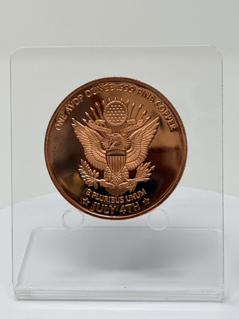Reverse of the 1 oz .999 copper round showing the Great Seal eagle, E Pluribus Unum and July 4th, standing on a clear acrylic stand against a plain light background