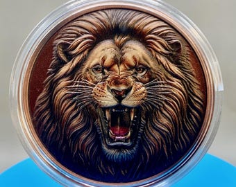 May include: A circular copper-toned medallion featuring a highly detailed, embossed relief of a roaring lion's head with a full mane. The lion is depicted with its mouth open, showing teeth and a dark interior, encased in a clear protective plastic capsule.