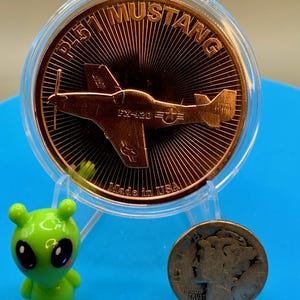 Front of a 1 oz .999 fine copper round with a P-51 Mustang design, standing in a display holder next to a small green alien figurine and a worn silver dime for size comparison, on a blue background.
