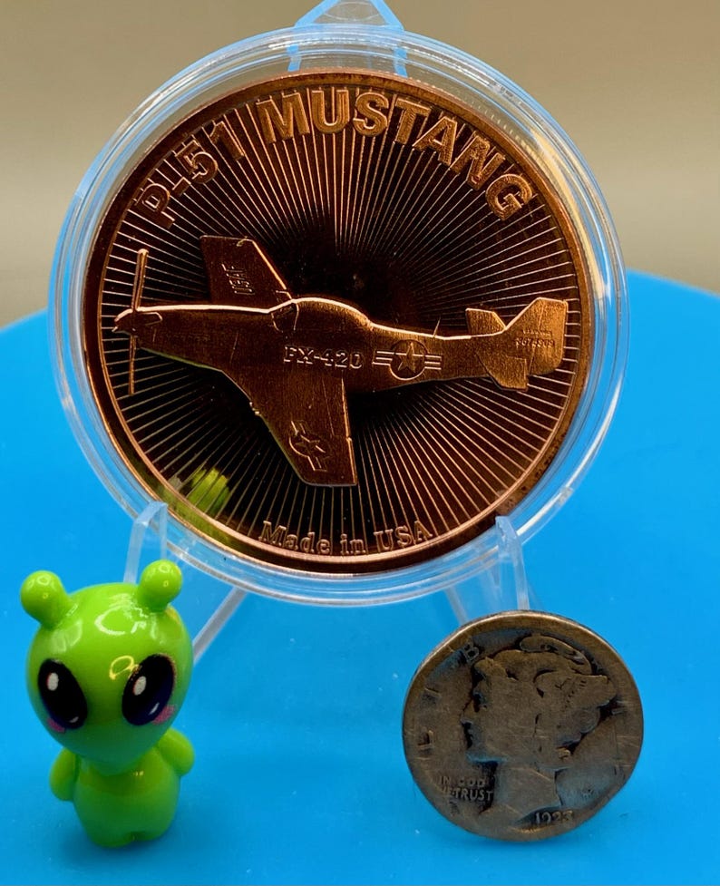 Front of a 1 oz .999 fine copper round with a P-51 Mustang design, standing in a display holder next to a small green alien figurine and a worn silver dime for size comparison, on a blue background.