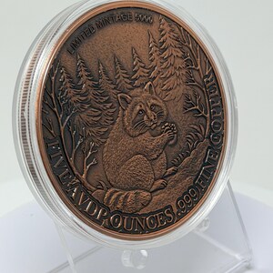 May include: A round copper coin encased in a clear plastic capsule, featuring a raccoon in a forest setting. The text reads LIMITED MINTAGE 5000 and FIVE AVD P OUNCES .999 FINE COPPER. The coin is displayed on a clear plastic stand.