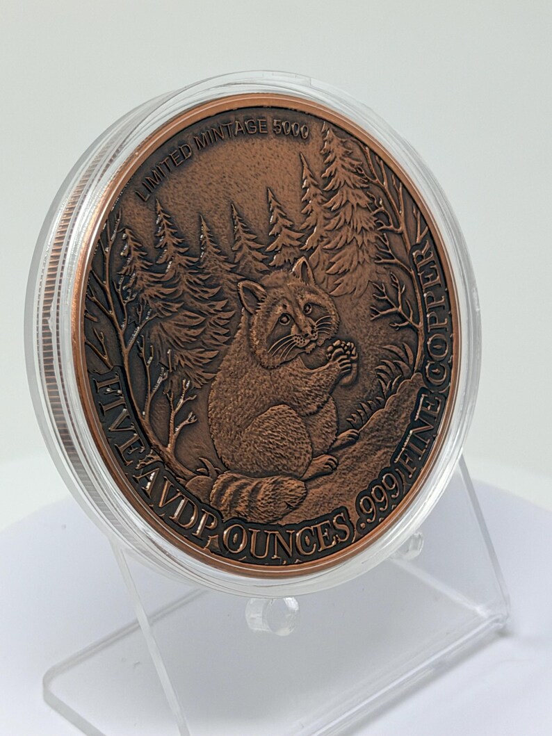 May include: A round copper coin encased in a clear plastic capsule, featuring a raccoon in a forest setting. The text reads LIMITED MINTAGE 5000 and FIVE AVD P OUNCES .999 FINE COPPER. The coin is displayed on a clear plastic stand.