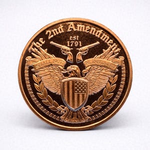 May include: A bronze-coloured coin featuring an eagle with outstretched wings, a shield with the American flag, and two crossed pistols. The text "The 2nd Amendment est 1791" and "SHALL NOT BE INFRINGED" are inscribed on the coin.