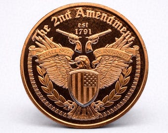 Peut inclure: Une pièce de monnaie de couleur bronze représentant un aigle aux ailes déployées, un bouclier avec le drapeau américain et deux pistolets croisés. Le texte "The 2nd Amendment est 1791" et "SHALL NOT BE INFRINGED" est inscrit.