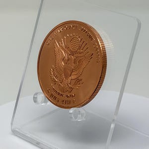 Angled rear view of the 1 oz copper round showing the Great Seal eagle reverse, tilted on a clear acrylic stand