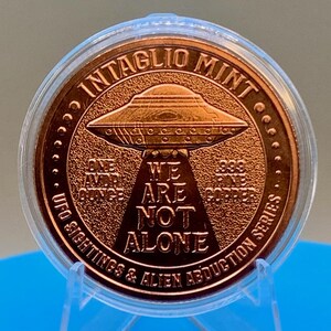 Reverse of the Intaglio Mint 1 oz .999 copper round showing a flying saucer projecting a tractor beam with the words WE ARE NOT ALONE, surrounded by the legend UFO SIGHTINGS & ALIEN ABDUCTION SERIES and the text ONE AVDP OUNCE .999 FINE COPPER, in a clear capsule.
