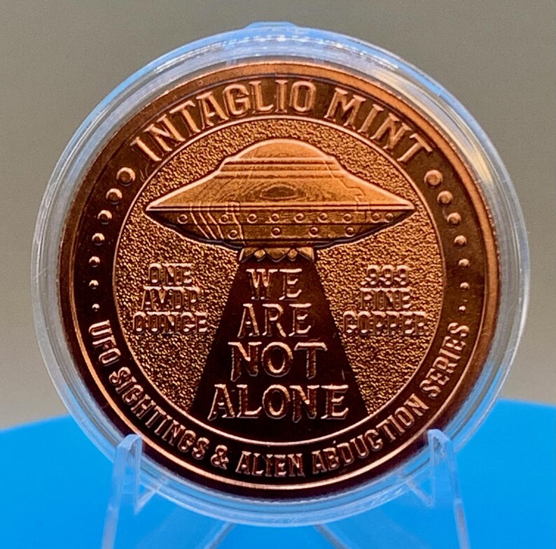 Reverse of the Intaglio Mint 1 oz .999 copper round showing a flying saucer projecting a tractor beam with the words WE ARE NOT ALONE, surrounded by the legend UFO SIGHTINGS & ALIEN ABDUCTION SERIES and the text ONE AVDP OUNCE .999 FINE COPPER, in a clear capsule.
