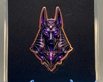 May include: A metal enamel pin featuring the head of Anubis with pointed ears, rendered in deep purple and black tones with copper-coloured metallic outlines. The pin is displayed inside a clear square protective case with black foam backing.