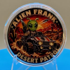 Alien Frank Desert Patrol 1 oz copper round in a clear capsule on a blue stand, showing the colorized alien driving a desert buggy with "Alien Frank" above and "Desert Patrol" below.