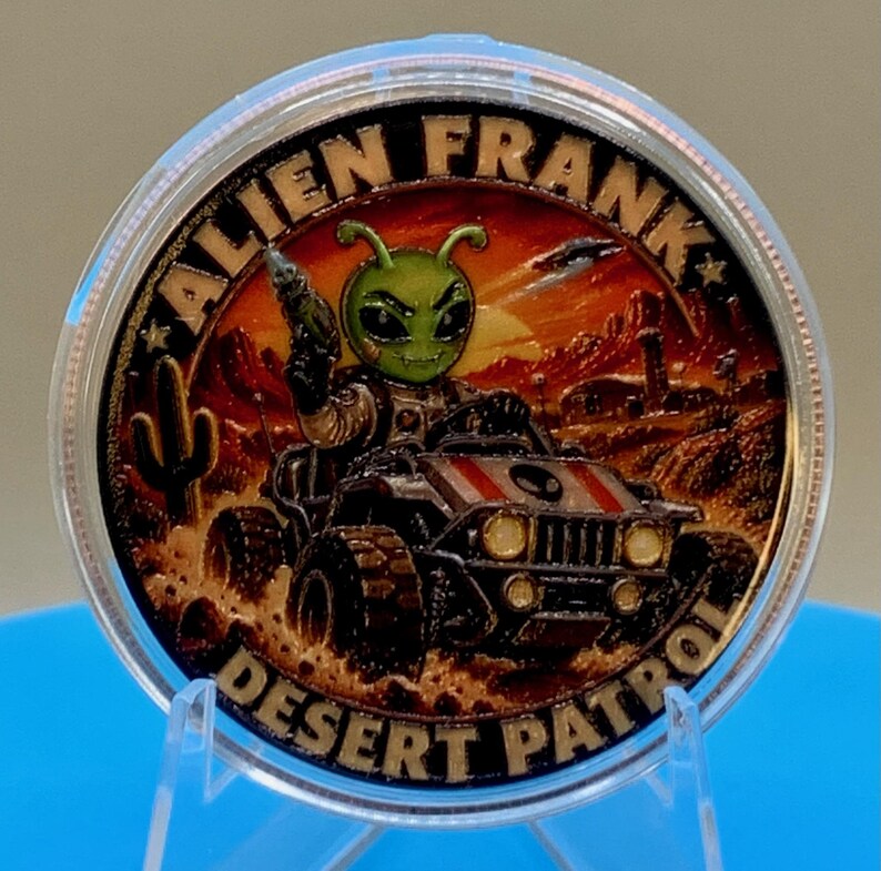 Alien Frank Desert Patrol 1 oz copper round in a clear capsule on a blue stand, showing the colorized alien driving a desert buggy with "Alien Frank" above and "Desert Patrol" below.