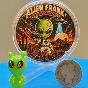 First Contact 1 oz copper round standing in its capsule beside a small green alien figurine and a silver coin for size comparison