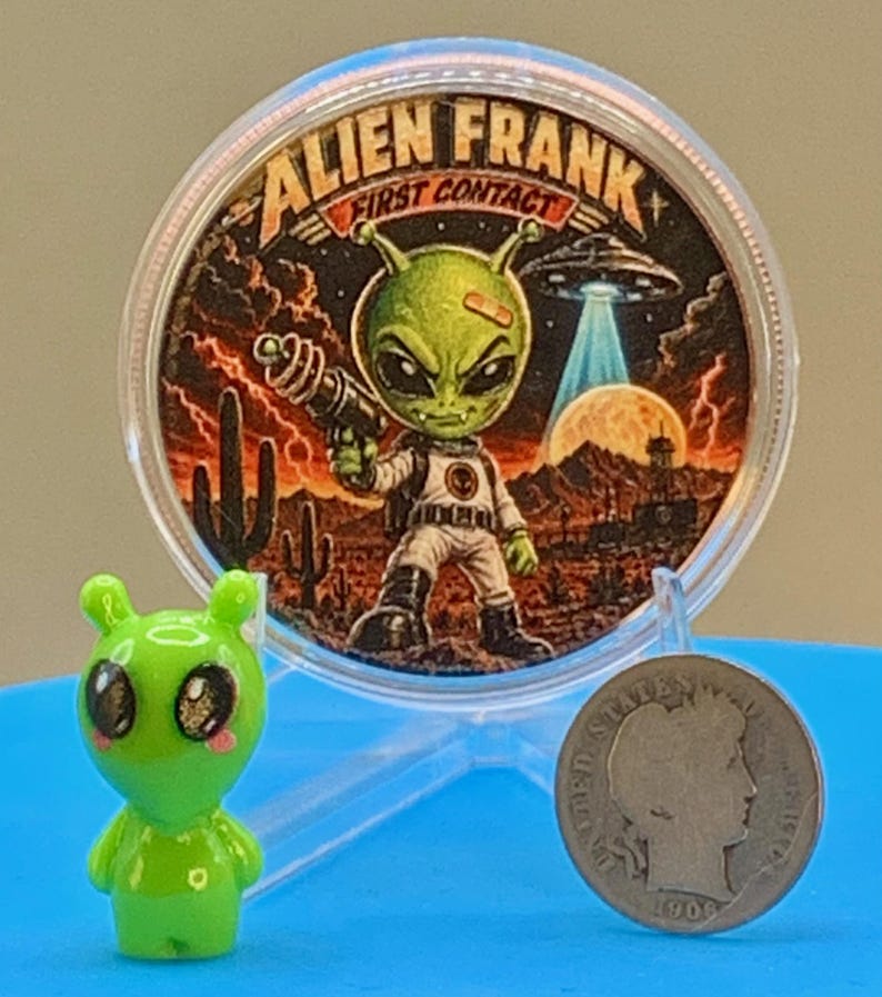 First Contact 1 oz copper round standing in its capsule beside a small green alien figurine and a silver coin for size comparison