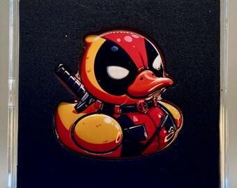 May include: A metal enamel pin shaped like a rubber duck dressed as the character Deadpool, featuring a red and yellow color scheme, black eye patches, a red beak, and a black sword handle protruding from the back, encased in a clear square case.