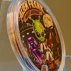 Angled view of the Night Arrival 1 oz copper round in its capsule showing the colorized green alien and purple UFO face and the rim of the coin