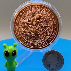 以下が含まれることがあります： FORTUNA FAVET、ONE AVDP OUNCE、.999 FINE COPPER、GOOD LUCK CHARMS、INTAGLIO MINTの文字が刻まれた、透明なプラスチックケース入りの銅色の丸いコイン。小さな緑色のエイリアンのフィギュアと、1906年製の銀色の米国コインが近くに置かれています。