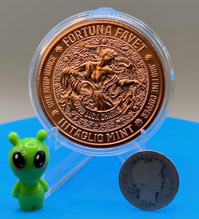 以下が含まれることがあります： FORTUNA FAVET、ONE AVDP OUNCE、.999 FINE COPPER、GOOD LUCK CHARMS、INTAGLIO MINTの文字が刻まれた、透明なプラスチックケース入りの銅色の丸いコイン。小さな緑色のエイリアンのフィギュアと、1906年製の銀色の米国コインが近くに置かれています。