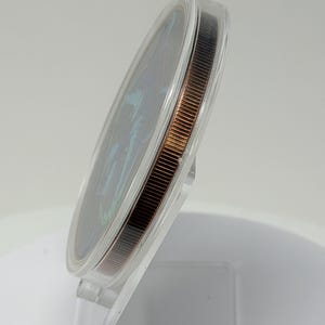 May include: A circular coin with a dark, iridescent face featuring blue and green highlights, encased in a clear, protective plastic capsule. The coin has a reeded, copper-toned edge and is displayed at an angle on a transparent stand.