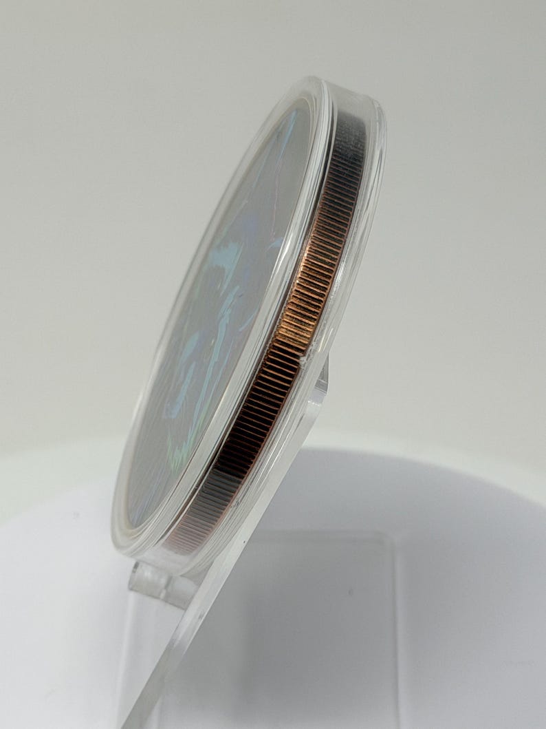 May include: A circular coin with a dark, iridescent face featuring blue and green highlights, encased in a clear, protective plastic capsule. The coin has a reeded, copper-toned edge and is displayed at an angle on a transparent stand.