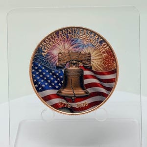 Front of a colorized 1 oz .999 copper round with the Liberty Bell, American flag and fireworks design, standing upright on a clear acrylic display stand against a plain light background