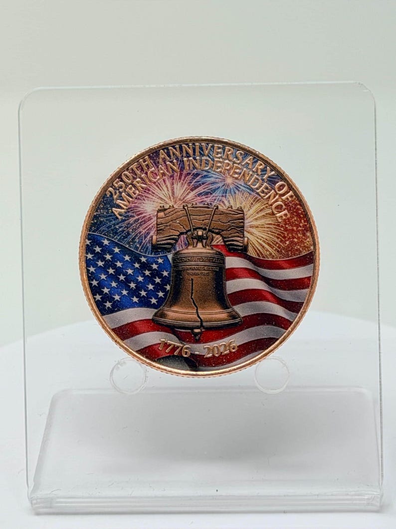 Front of a colorized 1 oz .999 copper round with the Liberty Bell, American flag and fireworks design, standing upright on a clear acrylic display stand against a plain light background