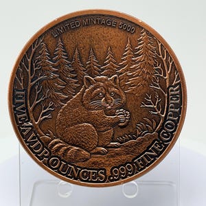 May include: A round copper coin featuring a raccoon sitting in a forest setting. The text reads LIMITED MINTAGE 5000 and FIVE AVP OUNCES .999 FINE COPPER. The design includes detailed pine trees and the raccoon holding a small object in its paws.