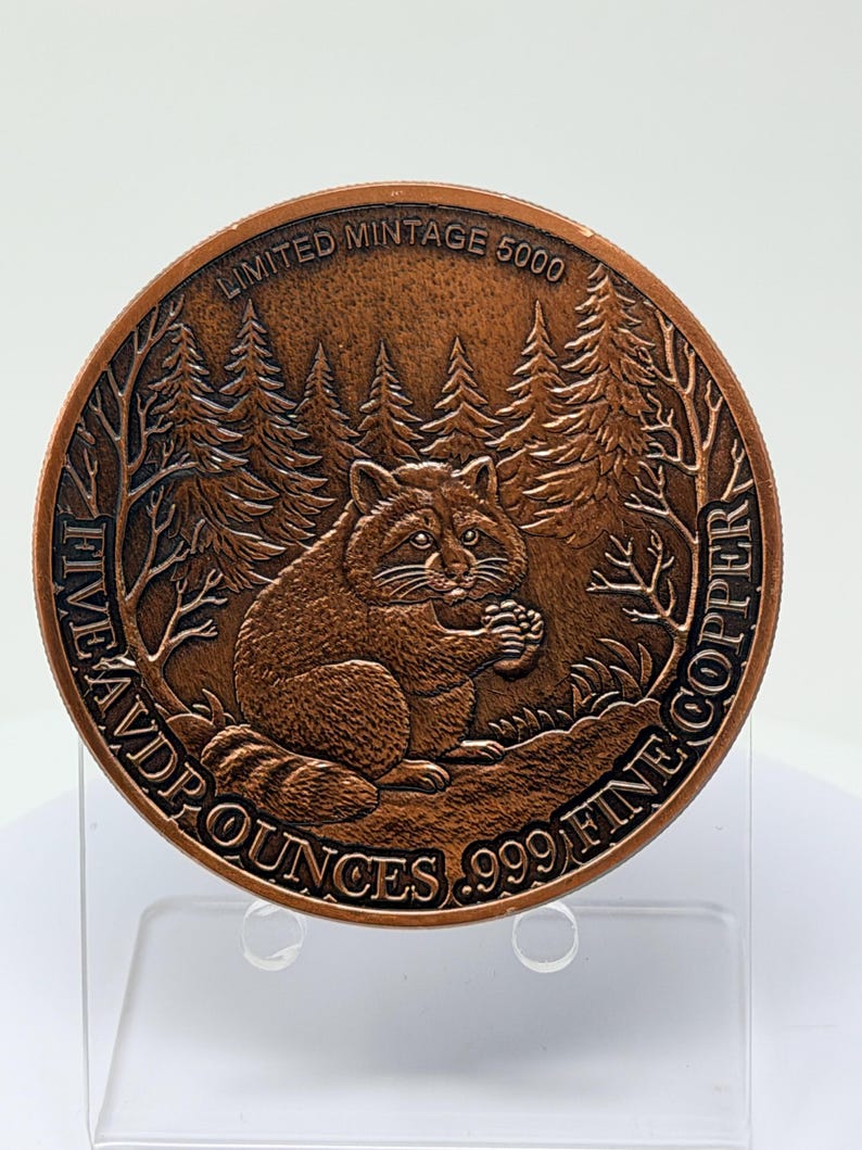 May include: A round copper coin featuring a raccoon sitting in a forest setting. The text reads LIMITED MINTAGE 5000 and FIVE AVP OUNCES .999 FINE COPPER. The design includes detailed pine trees and the raccoon holding a small object in its paws.