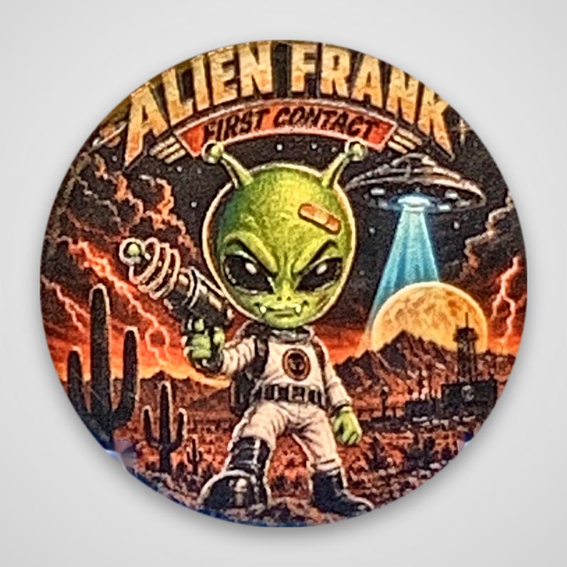 May include: A circular pinback button featuring a green alien character in a white spacesuit holding a ray gun. The background shows a desert landscape with cacti, a full moon, and a flying saucer. The text reads ALIEN FRANK FIRST CONTACT.