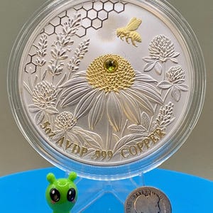 Reverse side of the Harmony Bee copper round in its capsule, featuring a gilded bee over honeycomb and coneflower engraving with rim text reading 5oz AVDP .999 COPPER, shown with a toy figurine and dime for scale.