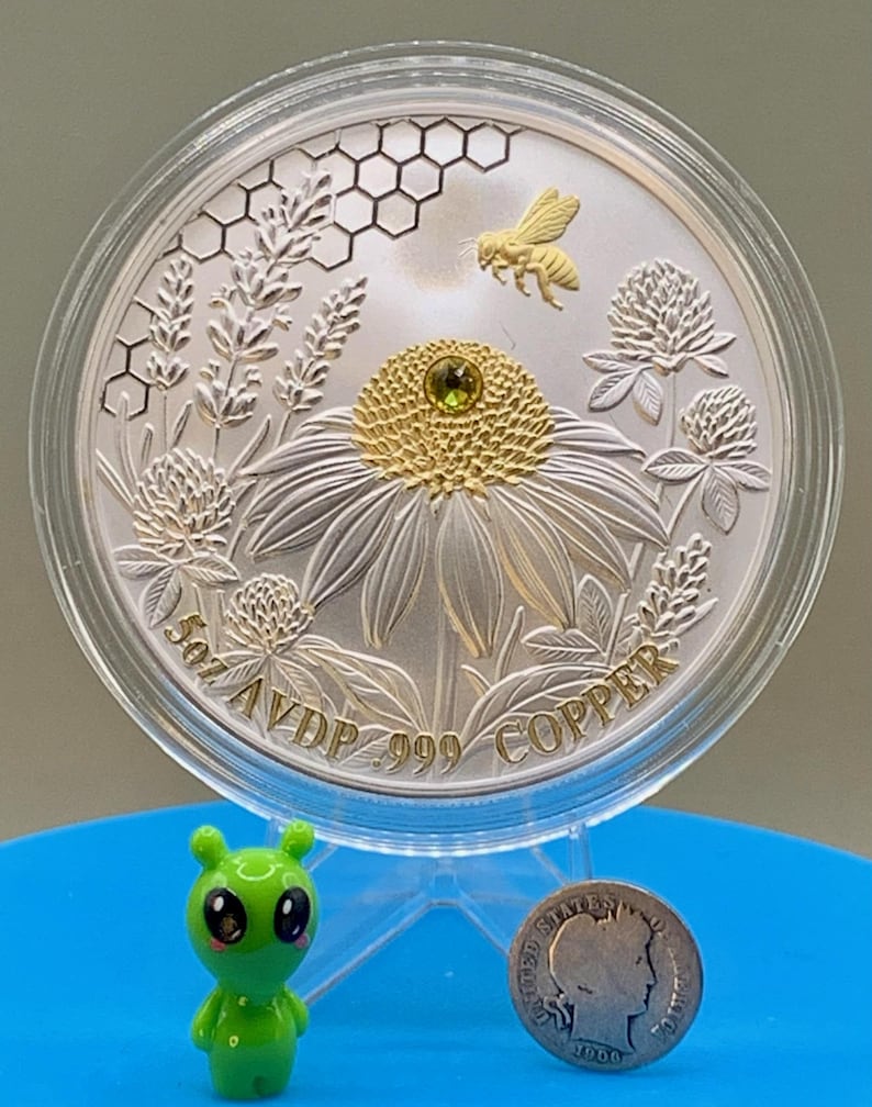 Reverse side of the Harmony Bee copper round in its capsule, featuring a gilded bee over honeycomb and coneflower engraving with rim text reading 5oz AVDP .999 COPPER, shown with a toy figurine and dime for scale.