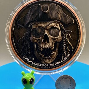 Puede incluir: Una moneda redonda de cobre con el relieve de una calavera que lleva un sombrero de pirata y un parche en el ojo. El texto 5 AVDP OUNCES OF .999 FINE COPPER est&aacute; grabado en el borde inferior. La moneda est&aacute; en una funda protectora.