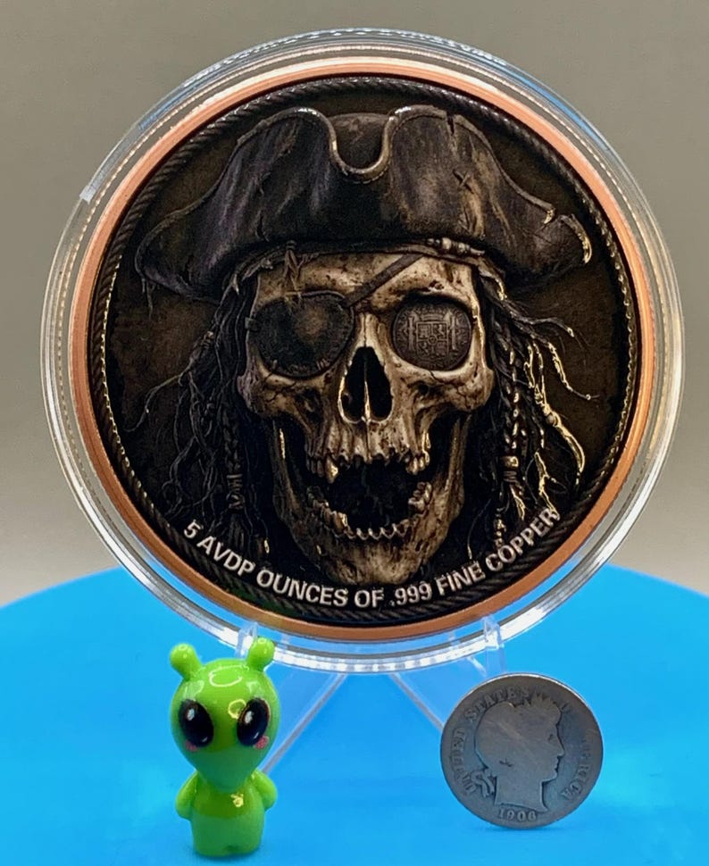 Puede incluir: Una moneda redonda de cobre con el relieve de una calavera que lleva un sombrero de pirata y un parche en el ojo. El texto 5 AVDP OUNCES OF .999 FINE COPPER est&aacute; grabado en el borde inferior. La moneda est&aacute; en una funda protectora.