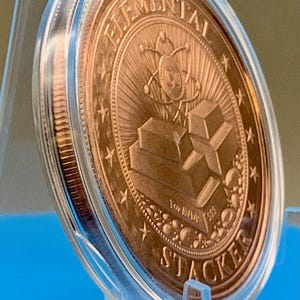 Side-angle view of the 1 oz copper round in a clear capsule on a blue stand, showing the reverse design with stacked copper bars and the thickness of the round.