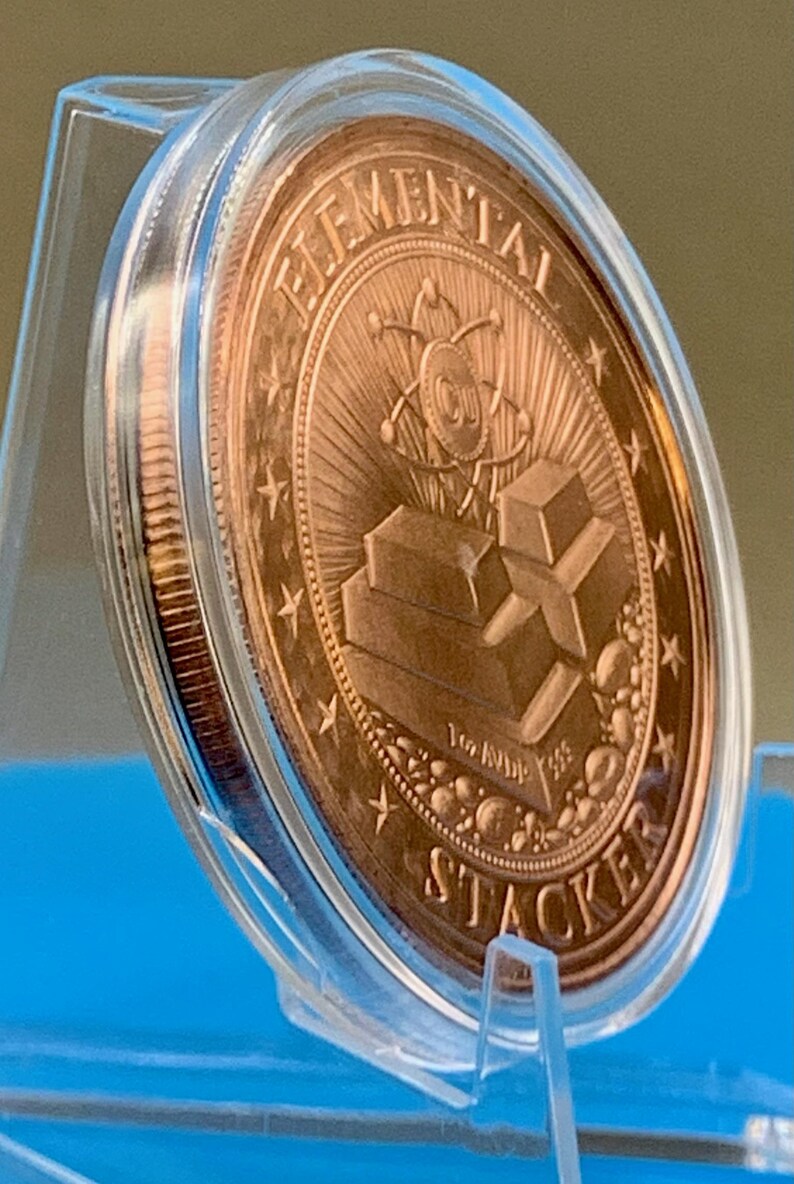 Side-angle view of the 1 oz copper round in a clear capsule on a blue stand, showing the reverse design with stacked copper bars and the thickness of the round.