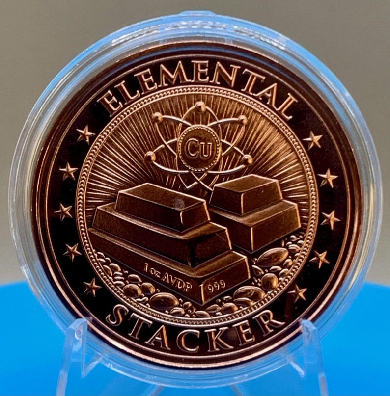 Close-up of the reverse of the 1 oz .999 fine copper round in a clear capsule, showing stacked copper bars with an atom and the Cu symbol, "Elemental" across the top and "Stacker" along the bottom, surrounded by a ring of stars.