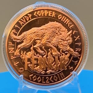 May include: A round copper coin encased in a clear plastic capsule. The coin features a relief of a wolf above flames with the text FENRIR, 1 AVP COPPER OUNCE, and TOOL2COIN, surrounded by a border of runic characters in a metallic copper finish.