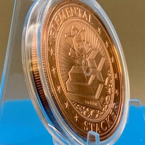 Side-angle view of the 1 oz copper round in a clear capsule on a blue stand, showing the reverse design with stacked copper bars and the thickness of the round.