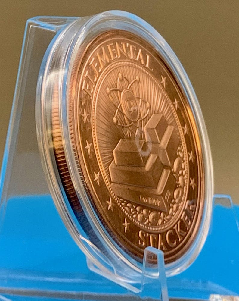 Side-angle view of the 1 oz copper round in a clear capsule on a blue stand, showing the reverse design with stacked copper bars and the thickness of the round.