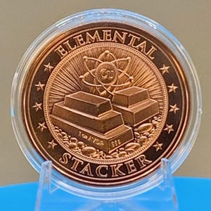 Reverse of the First Contact 1 oz copper round in its capsule: "Elemental Stacker" lettering around a stack of copper bars beneath an atom symbol, ringed by stars