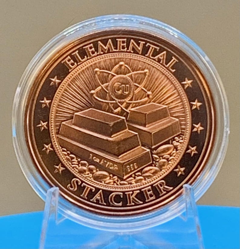 Reverse of the First Contact 1 oz copper round in its capsule: "Elemental Stacker" lettering around a stack of copper bars beneath an atom symbol, ringed by stars