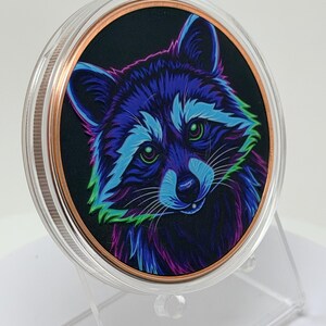 May include: A circular collectible coin featuring a vibrant, neon-colored illustration of a raccoon against a black background. The raccoon is rendered in shades of blue, purple, and green. The coin has a copper-toned rim and a clear protective casing.