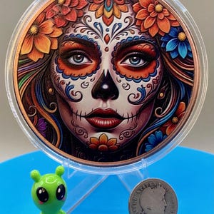 Collectible .999 fine copper legal tender coin featuring a vibrant sugar skull woman design inspired by Día de los Muertos traditions, shown for scale next to the shop's alien figurine and a silver dime