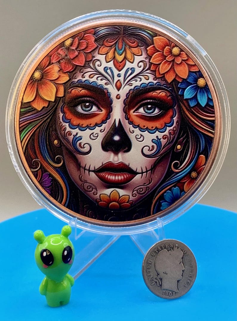 Collectible .999 fine copper legal tender coin featuring a vibrant sugar skull woman design inspired by Día de los Muertos traditions, shown for scale next to the shop's alien figurine and a silver dime