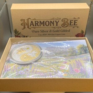 Overhead view of the open Harmony Bee copper round packaging, showing the round in its sealed capsule on top of a printed certificate card inside the box.