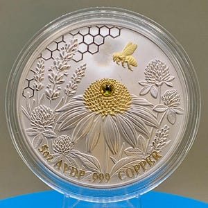 Straight-on view of the reverse side of the Harmony Bee copper round in its capsule, showing a gold bee, honeycomb, and coneflower engraving with rim text reading 5oz AVDP .999 COPPER.