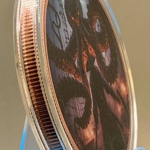 Puede incluir: Una moneda redonda de metal color cobre encerrada en una c&aacute;psula protectora de pl&aacute;stico transparente. La moneda presenta una ilustraci&oacute;n gr&aacute;fica detallada de un pulpo oscuro con tent&aacute;culos largos sobre un fondo de l&iacute;neas verticales.