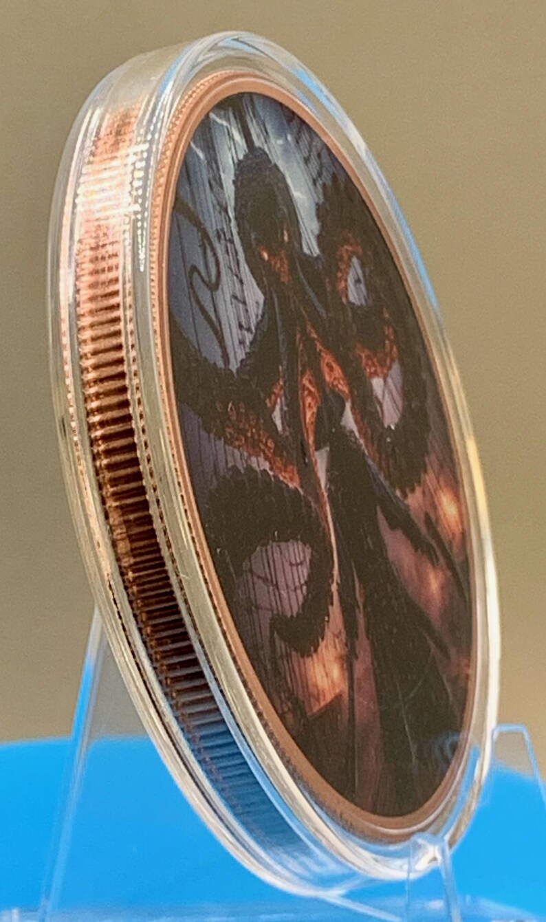 Puede incluir: Una moneda redonda de metal color cobre encerrada en una c&aacute;psula protectora de pl&aacute;stico transparente. La moneda presenta una ilustraci&oacute;n gr&aacute;fica detallada de un pulpo oscuro con tent&aacute;culos largos sobre un fondo de l&iacute;neas verticales.