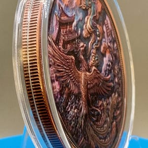 Angled side view of the copper Phoenix round with an iridescent patina finish in its protective capsule, showing the coin's edge against a blue background.