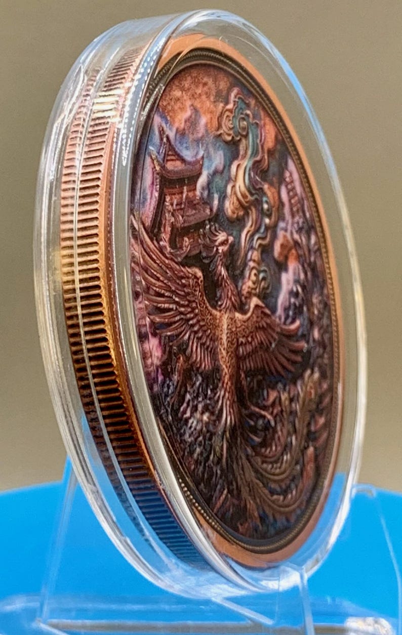 Angled side view of the copper Phoenix round with an iridescent patina finish in its protective capsule, showing the coin's edge against a blue background.