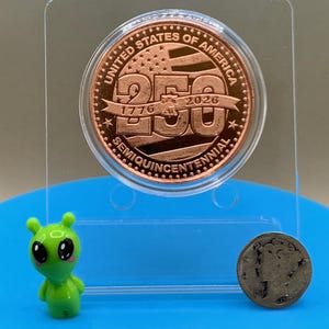 Close-up of United States Semiquincentennial copper round showing 1776 and 2026 dates, with small alien figurine for scale reference