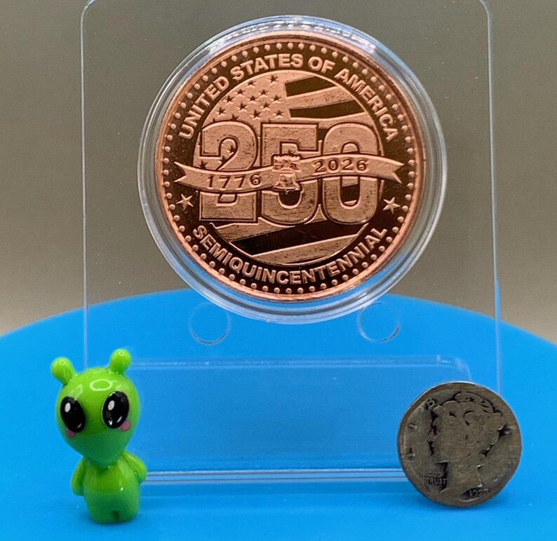 Close-up of United States Semiquincentennial copper round showing 1776 and 2026 dates, with small alien figurine for scale reference